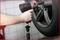 A hand guides an impact wrench to a car tyre to perform wheel mounting or dismounting. Workshop tiles are visible in the background.