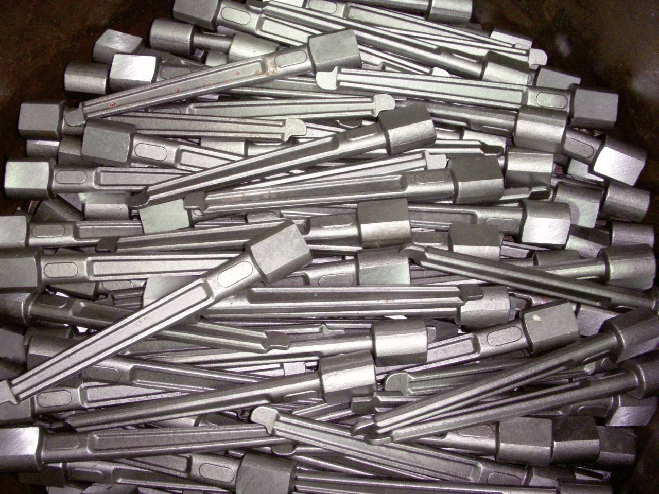 A stack of forged metal chisels with rectangular heads and long handles, arranged in a haphazard manner.