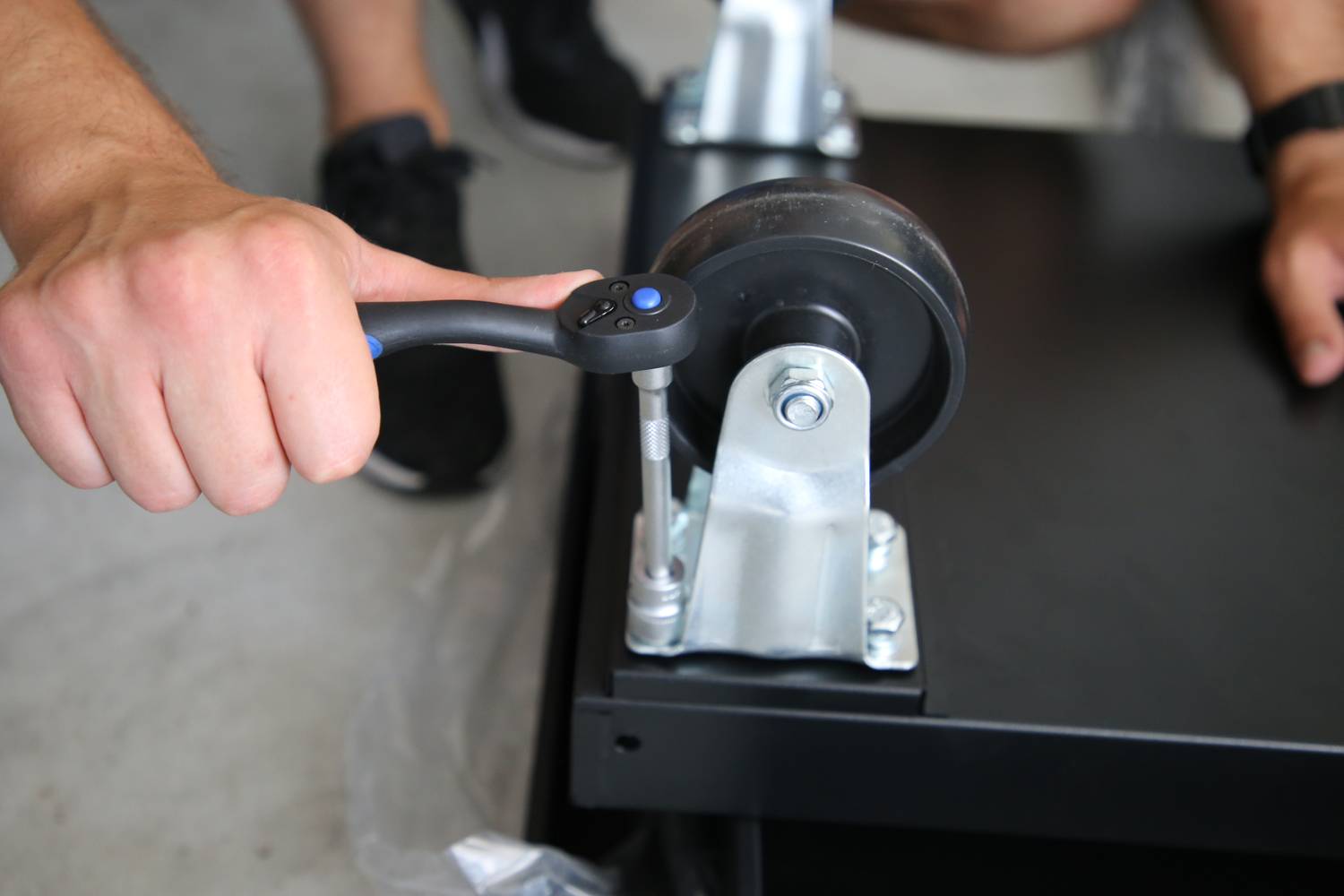 A person is tightening the screw of a swivel castor on a black piece of furniture with a spanner.