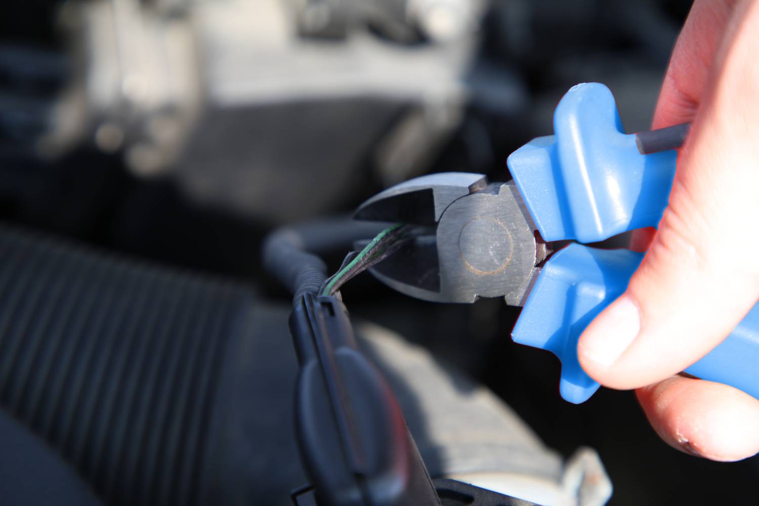 A hand is holding a blue side cutters and cutting a cable. The background is blurred, with the focus on the tool.