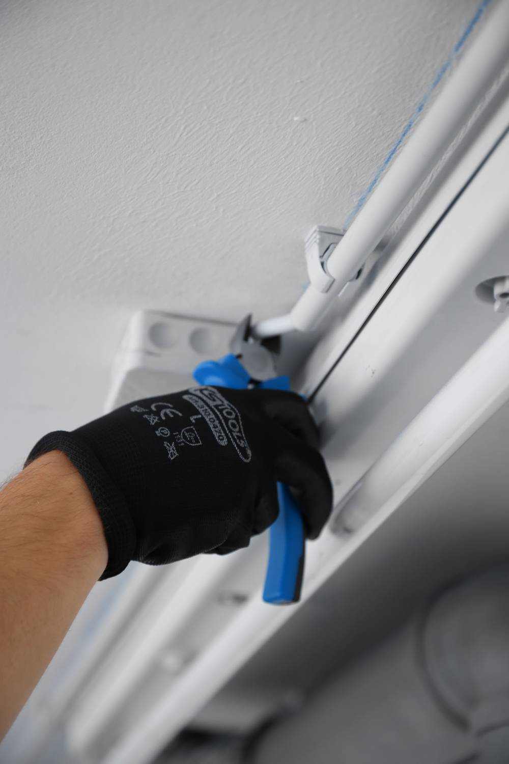 A person wearing black gloves is using a blue pair of pliers to secure a cable to a ceiling.
