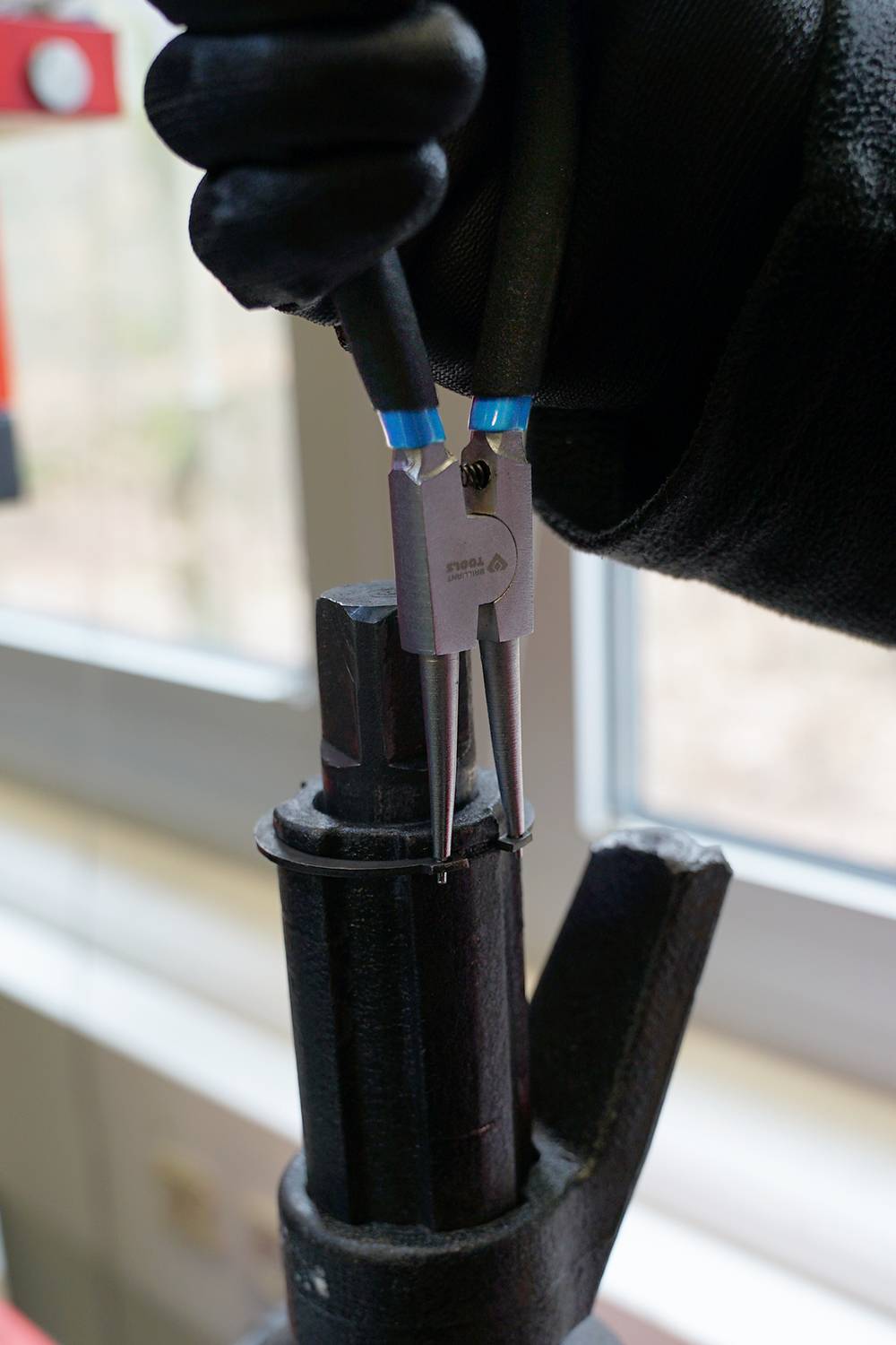 A person is using a pair of needle-nose pliers to remove a small black clip from a metallic object.