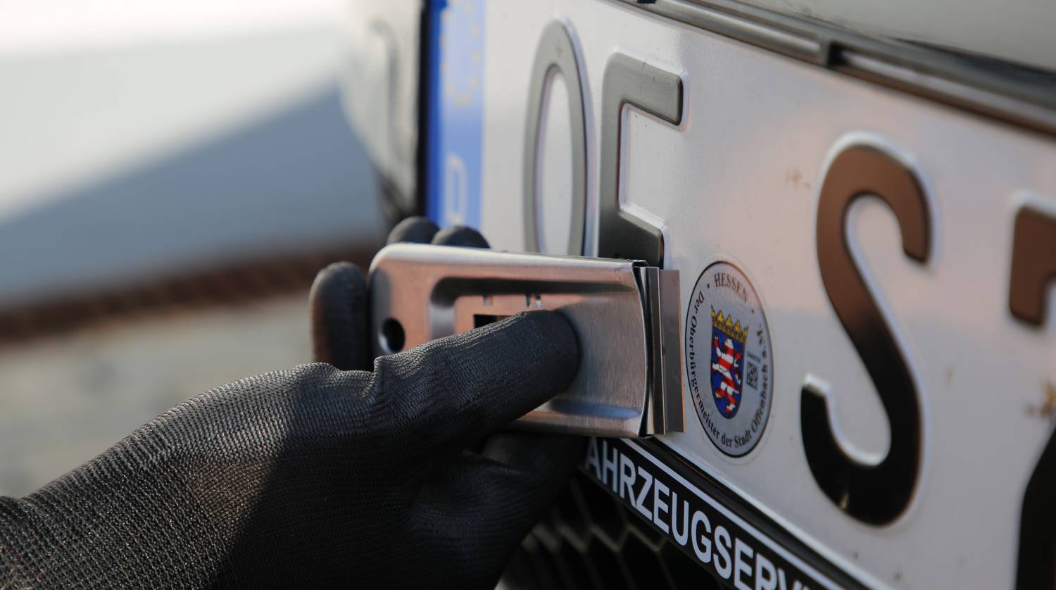 A person is attaching a German vehicle number plate with a tool. A seal is visible in the background.