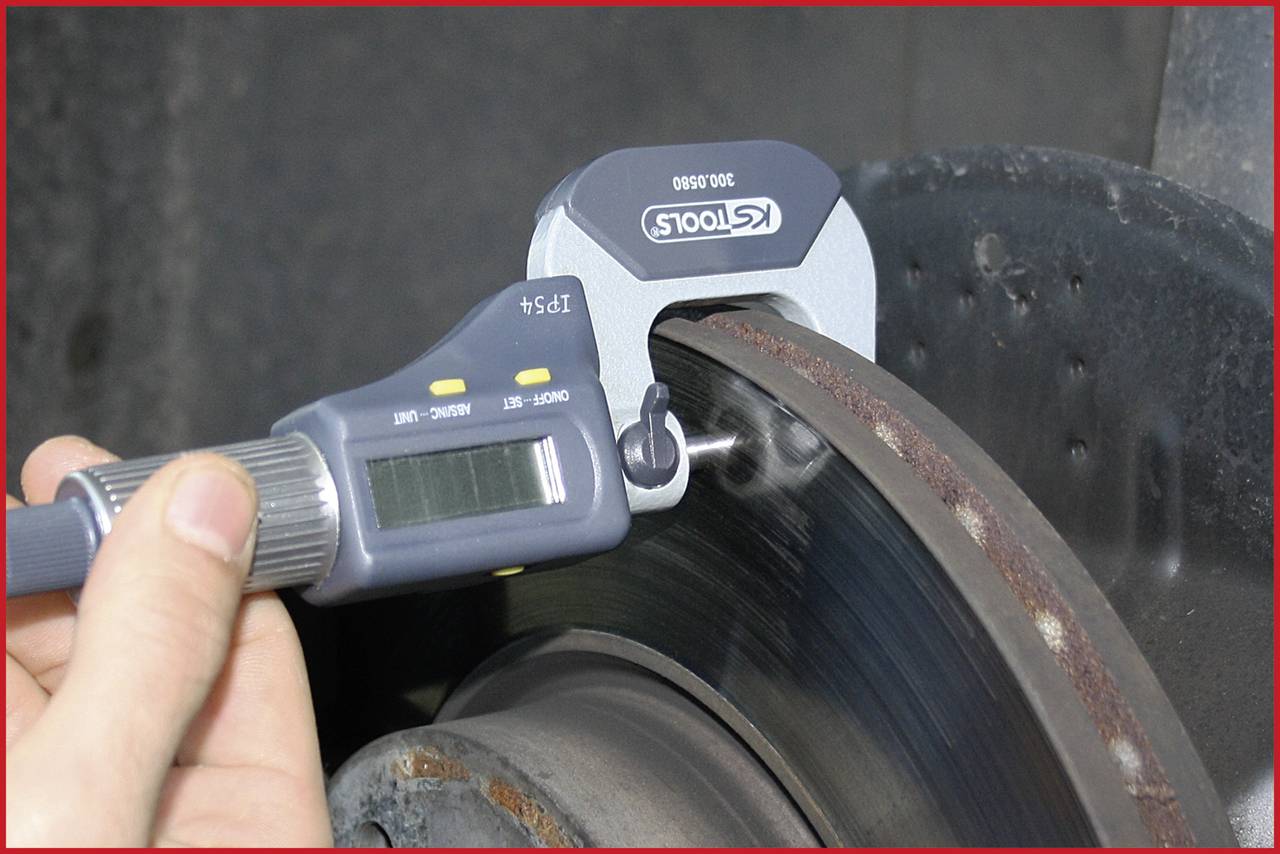 A hand is holding a digital calliper measuring the diameter of a brake disc. The display shows the measurement result.