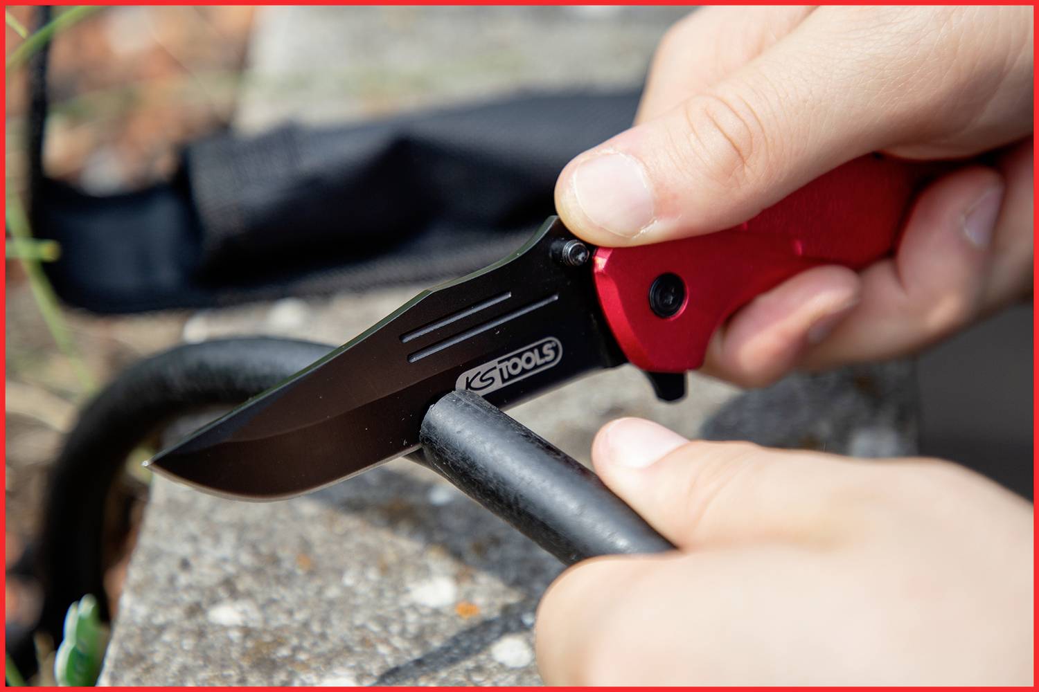 A person is cutting a black cable with a red folding knife on a stone surface. Close-up of hands and the knife.