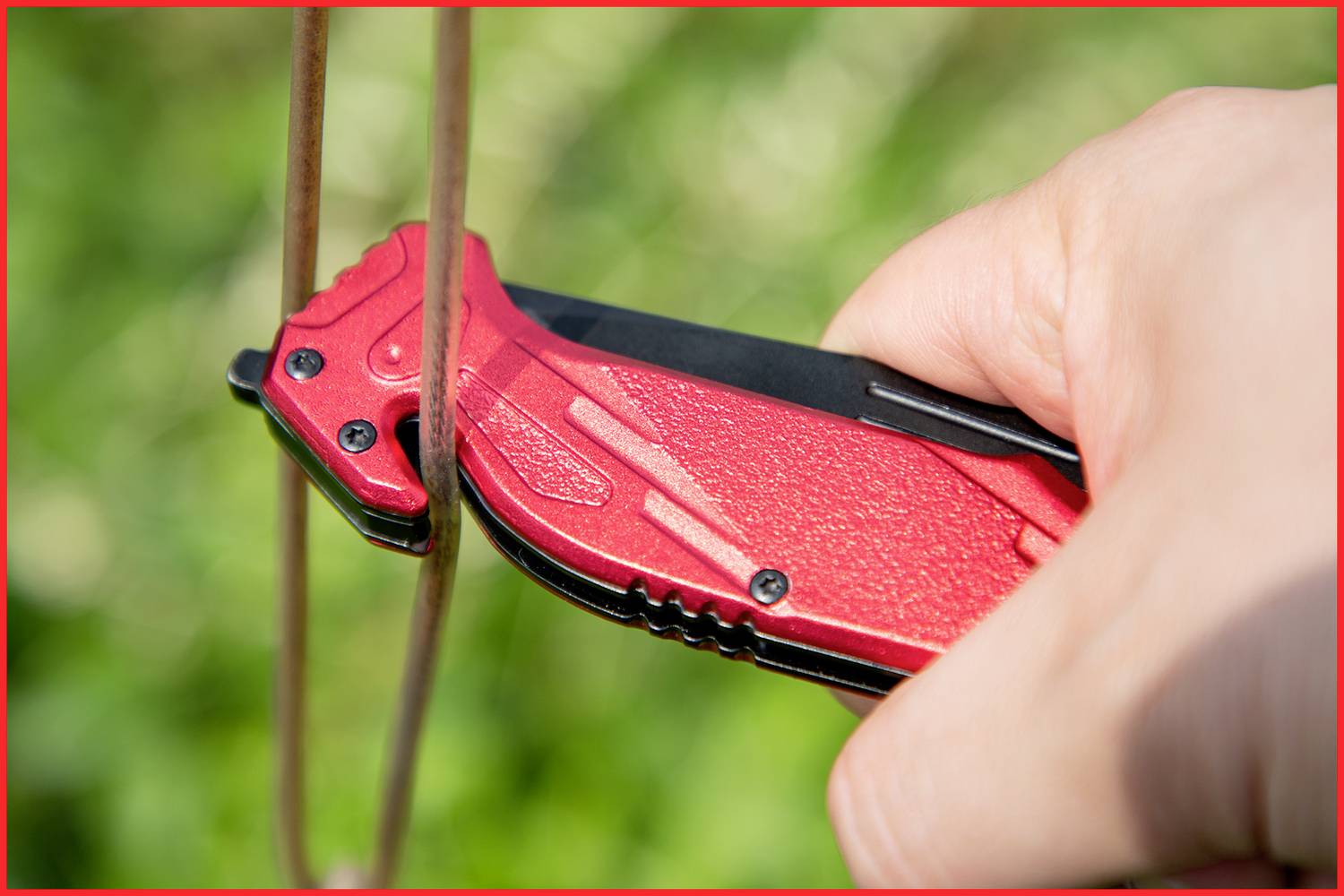 A hand holds a red folding knife cutting through a thin metal wire. Green background blurred.