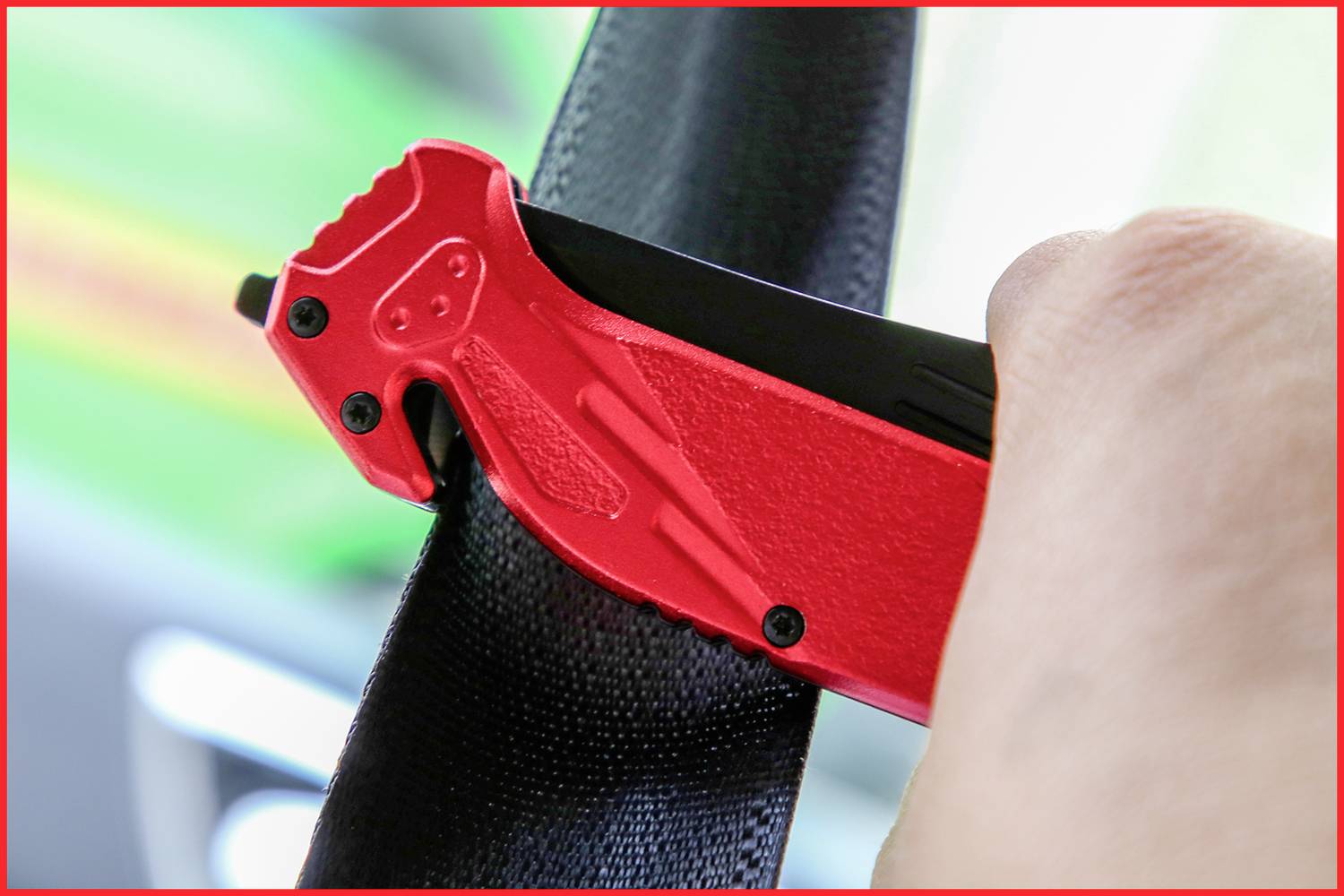 A hand is holding a red safety tool, about to cut through a black safety belt, with a blurred background.