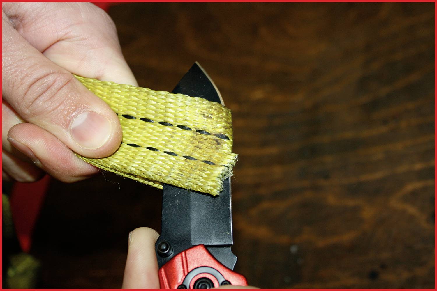 A hand cuts a yellow ribbon with two black lines using a red penknife. Background of dark wood.