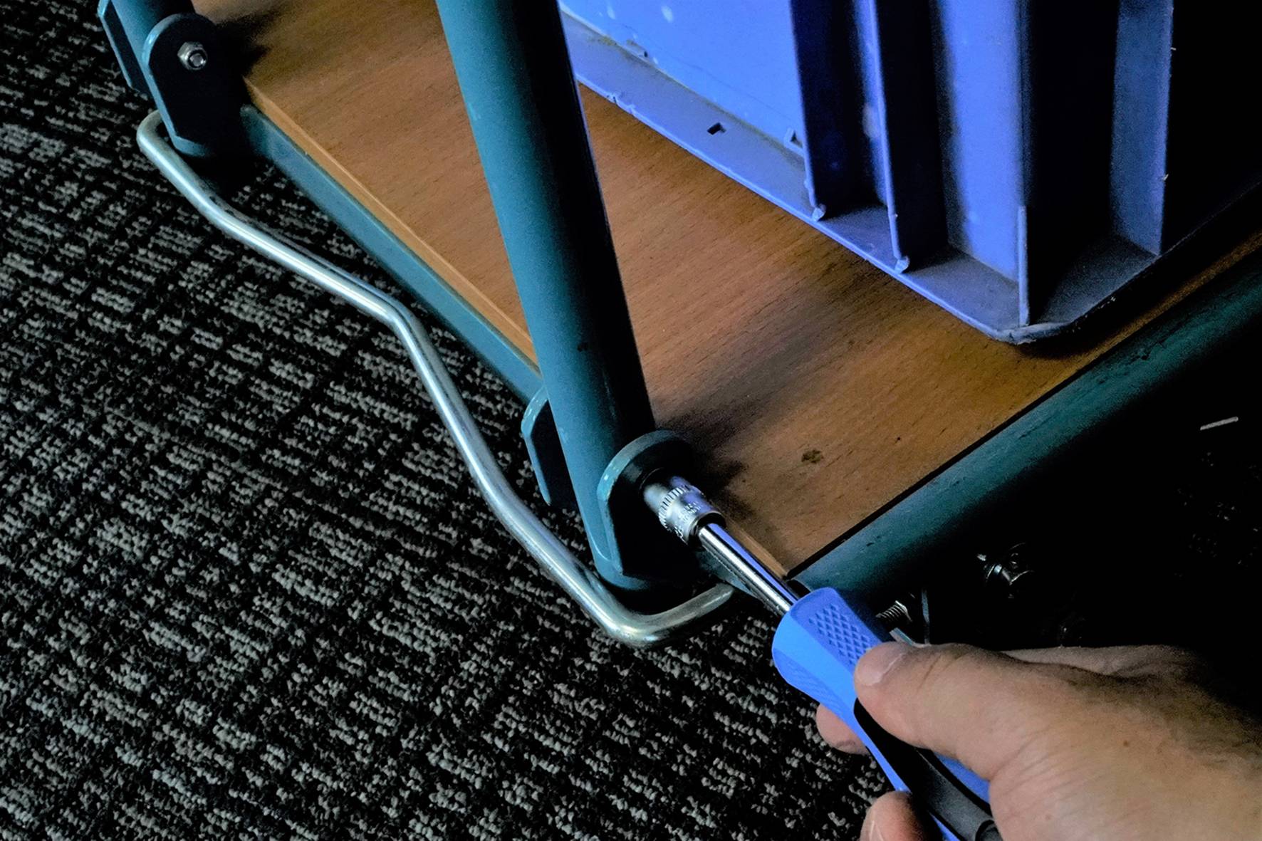 A person is tightening a screw on a metal frame with a spanner. A blue container is sitting on the frame.