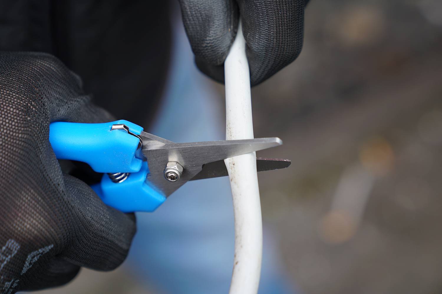 A person wearing black gloves is cutting a white cable with blue scissors.