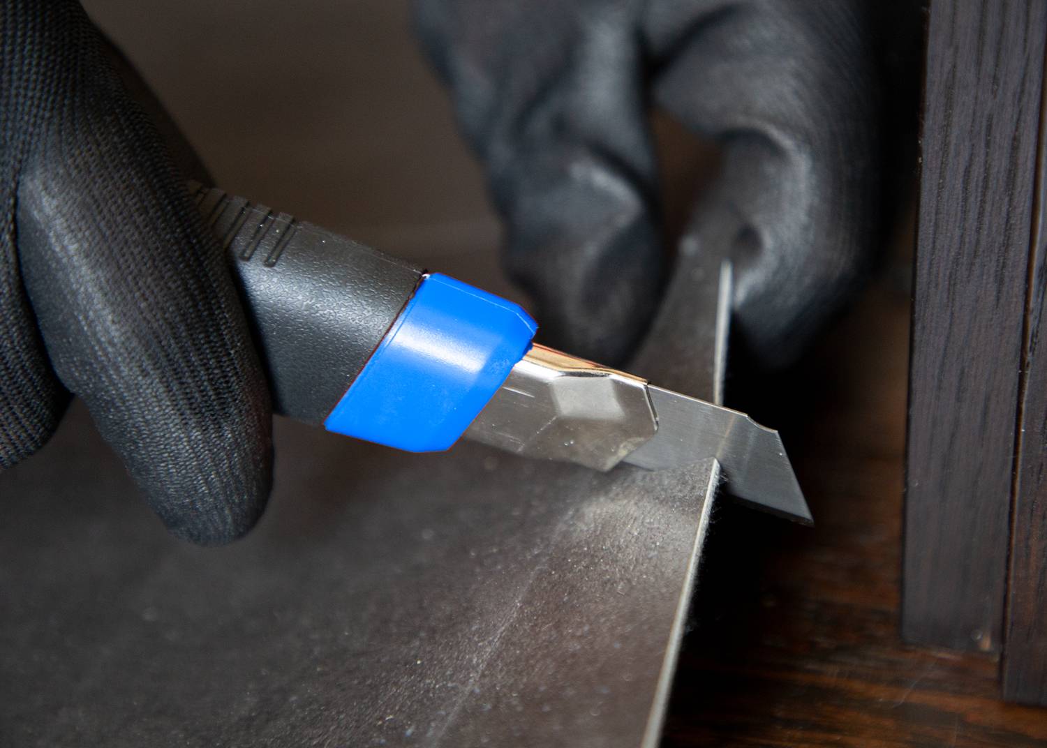 A person is cutting a piece of material with a blue craft knife. Hands wearing black gloves are holding the knife securely.
