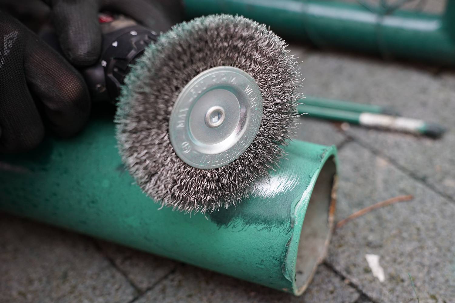 An angle grinder with a wire brush is working on a green metal pipe. The worker is wearing black protective gloves.