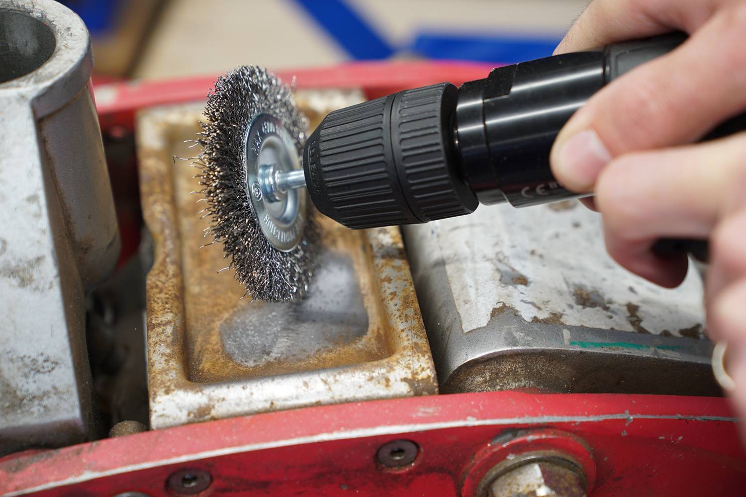 A hand holds an electric drill with a wire brush attachment, cleaning a rusty metal surface.