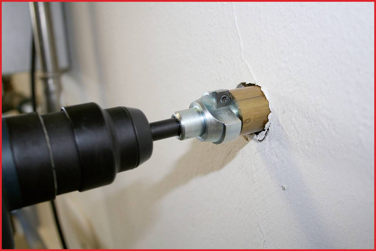 A drill with a drill bit is boring through a white wall to create a round hole. The tool is black and the drilling accessory is silver.