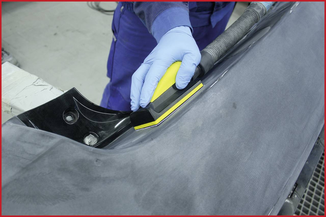 A person is wearing blue gloves and sanding a black car bumper with a yellow sanding pad.