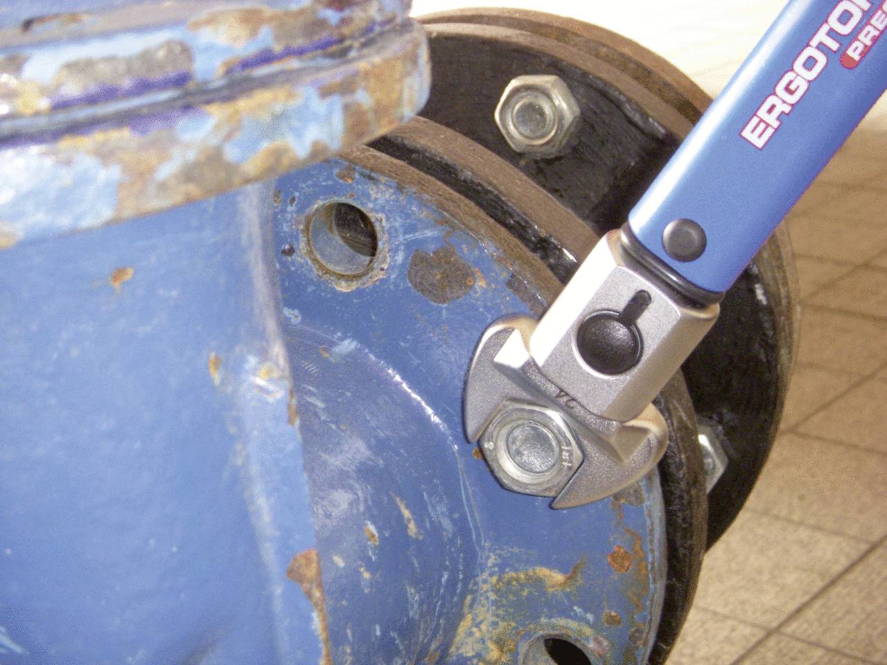 A spanner tightens a screw on a rusty, blue metal object. In the background, blurred metal parts can be seen.