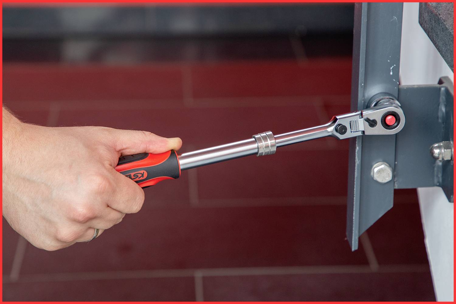 A hand is holding an adjustable torque wrench attached to a metal structure. The floor beneath is tiled in red.