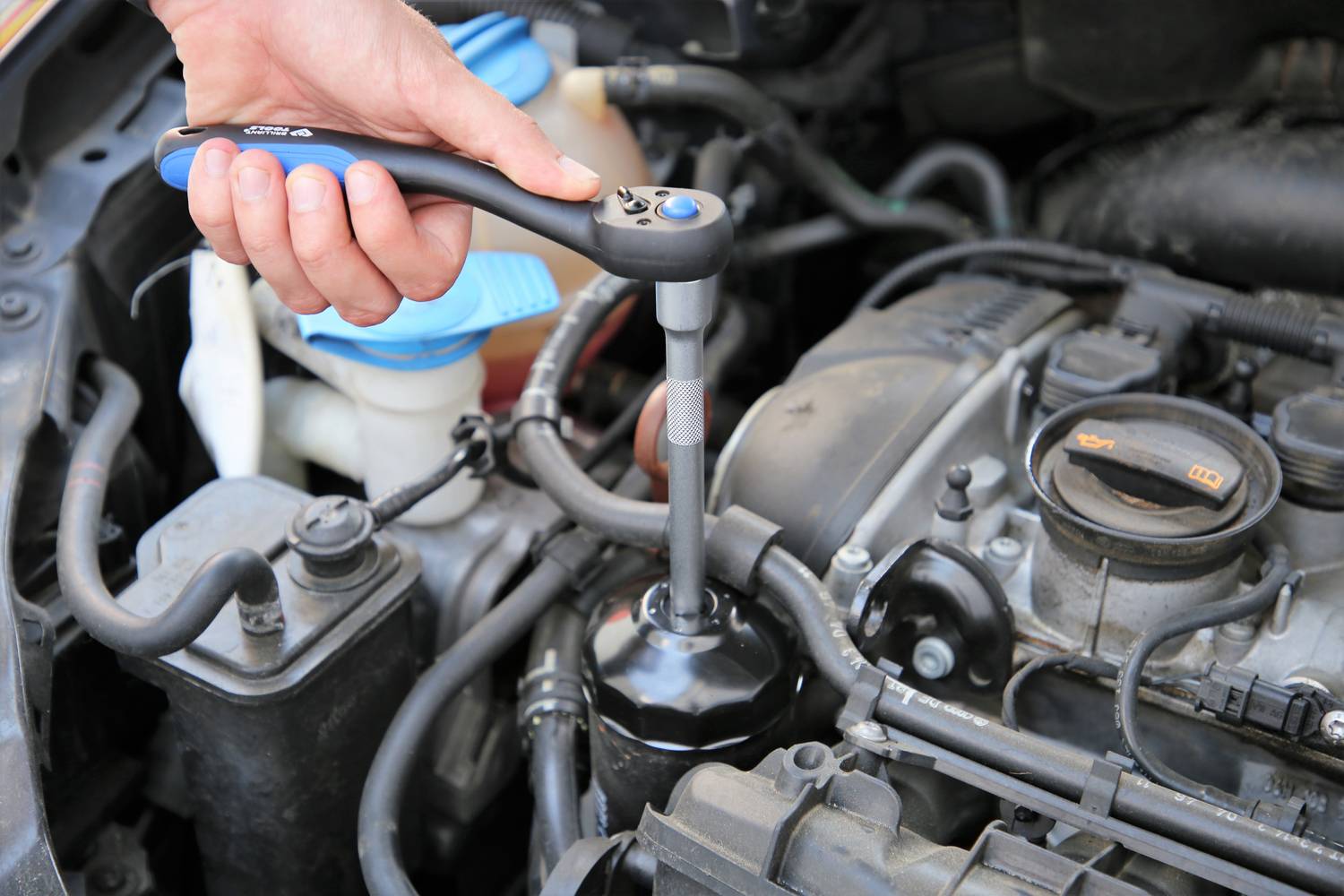 A hand is using a ratchet to work on an open car engine. Various engine components are visible.