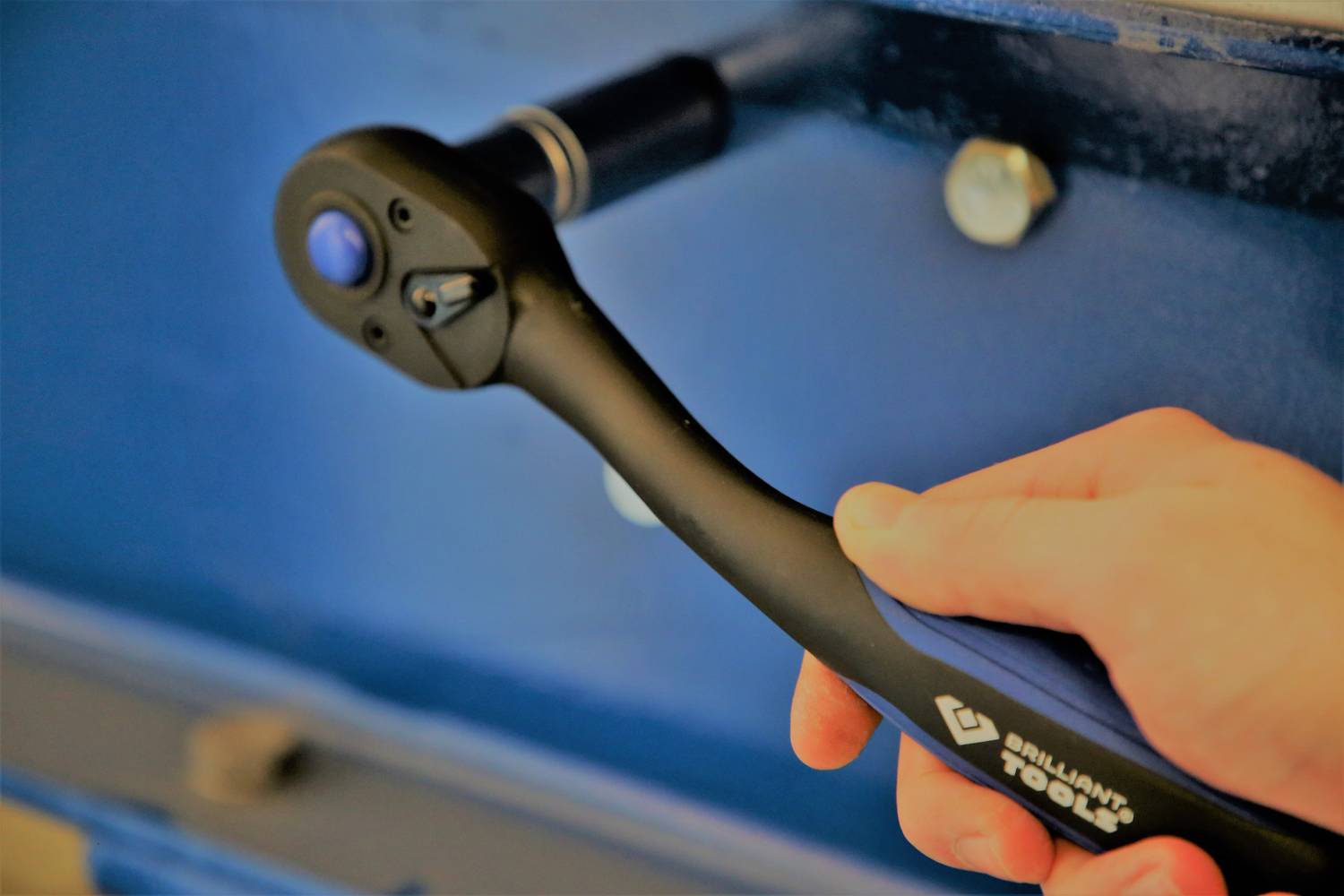 A hand holds a black ratchet spanner that is working on a blue metal surface.