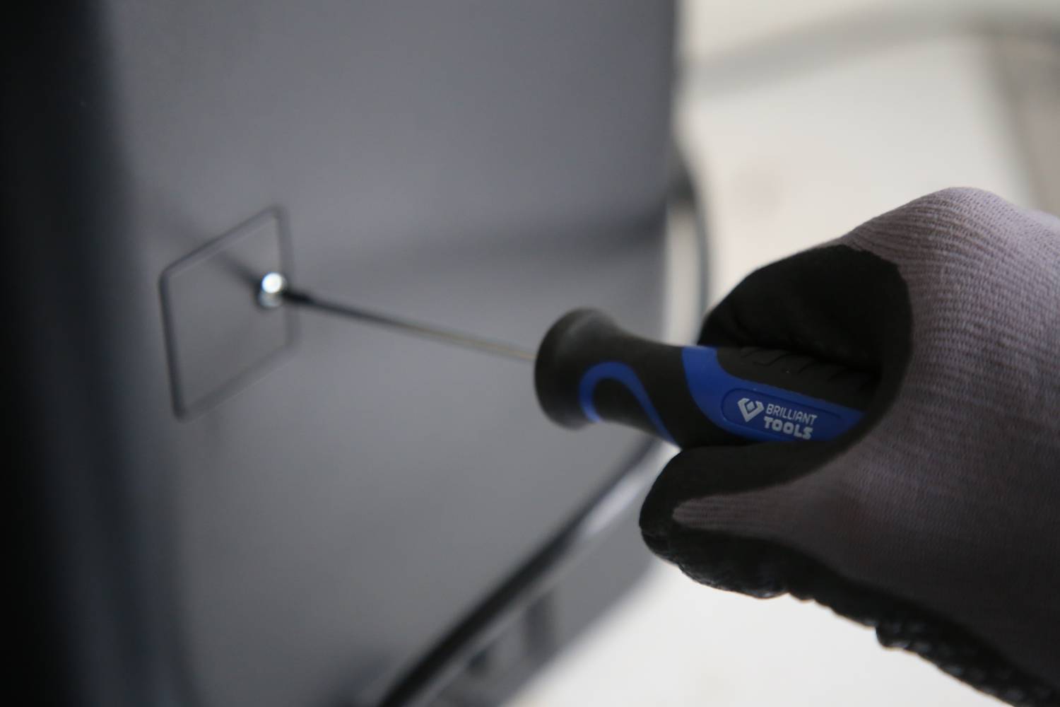 A gloved hand holds a screwdriver, turning it into the screw of a black device. Background blurred.