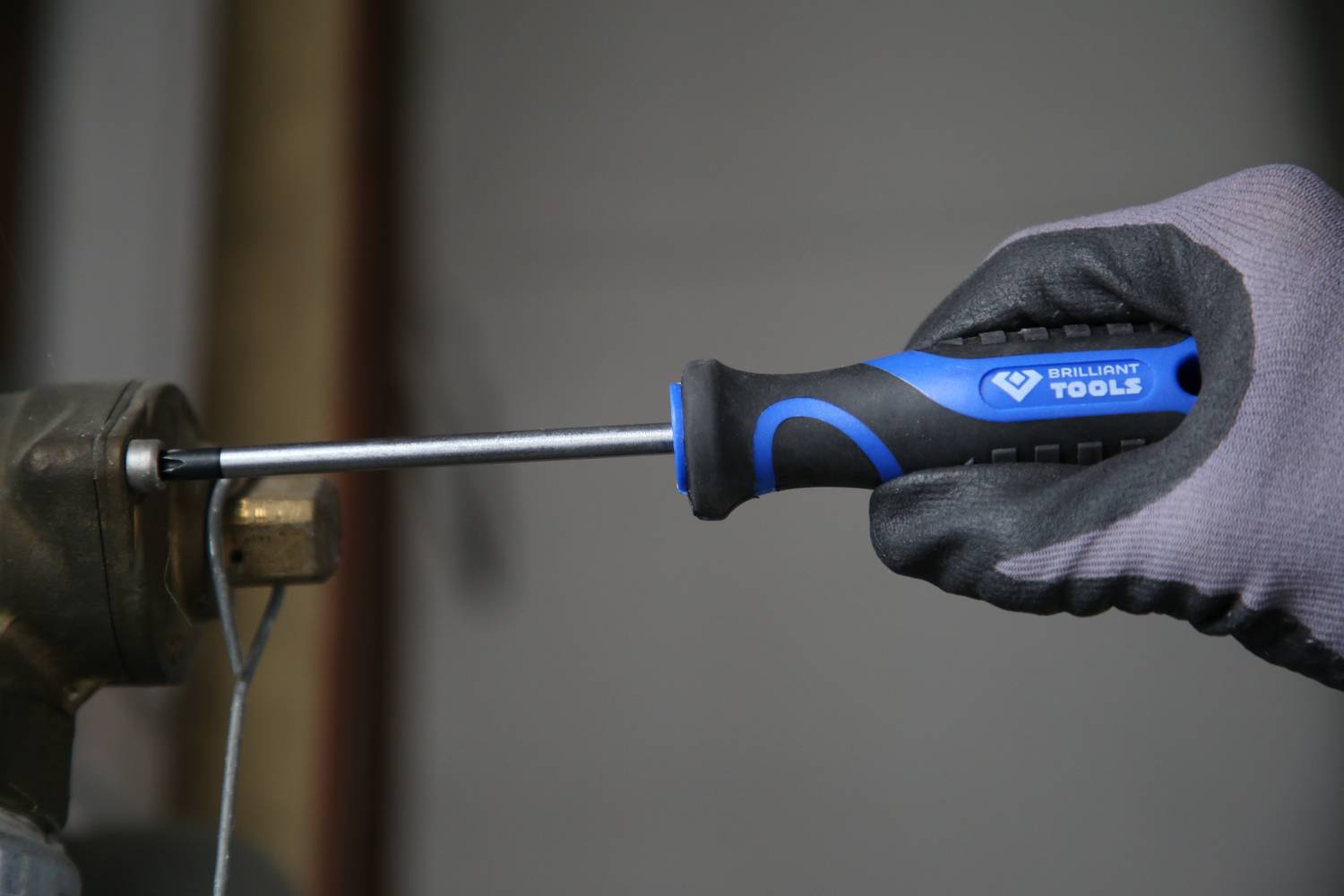 A hand wearing a glove holds a blue screwdriver and tightens a screw into a metal bracket.