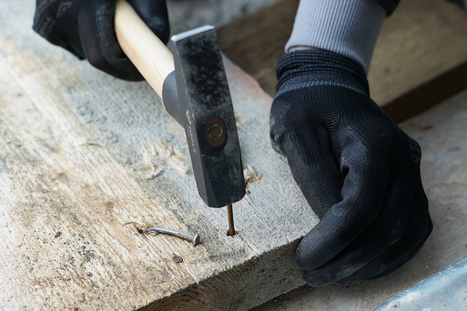 A person is hammering a nail into a wooden board, wearing black work gloves.