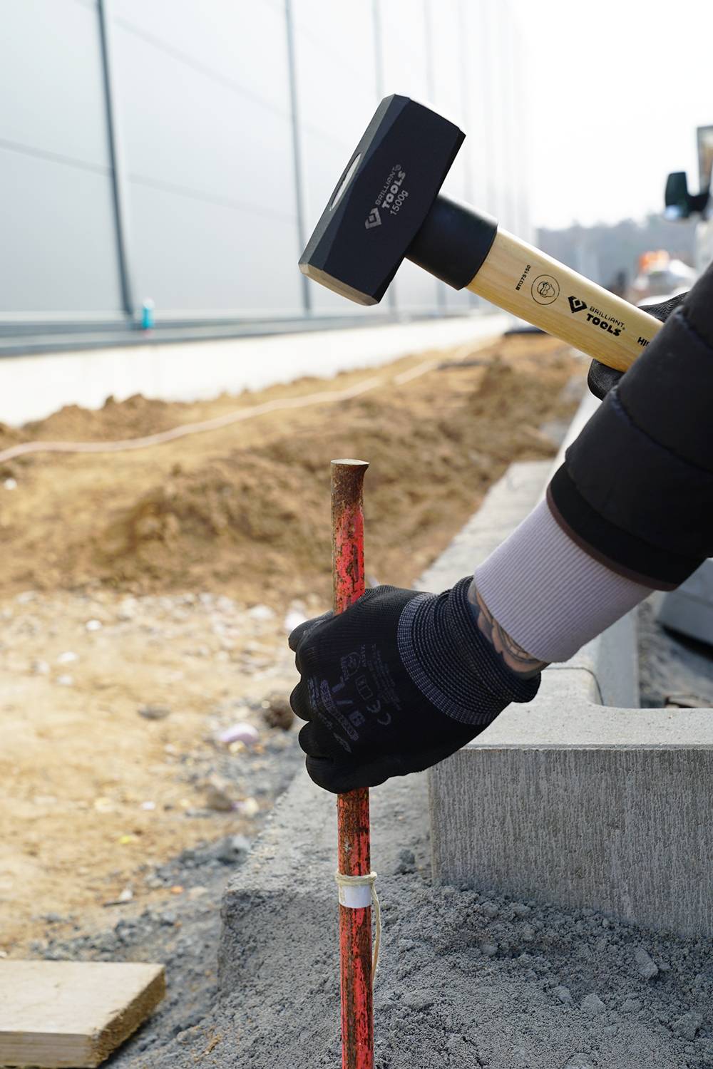 A person is hammering a wooden post into the ground. A construction site is visible in the background. A glove covers the person's hand.