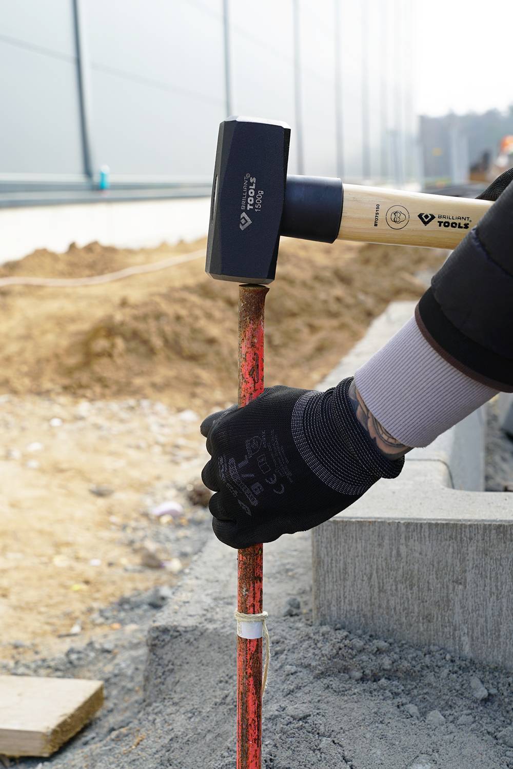 A person is hammering a red metal rod on a construction site. Soil and concrete blocks are visible in the background.