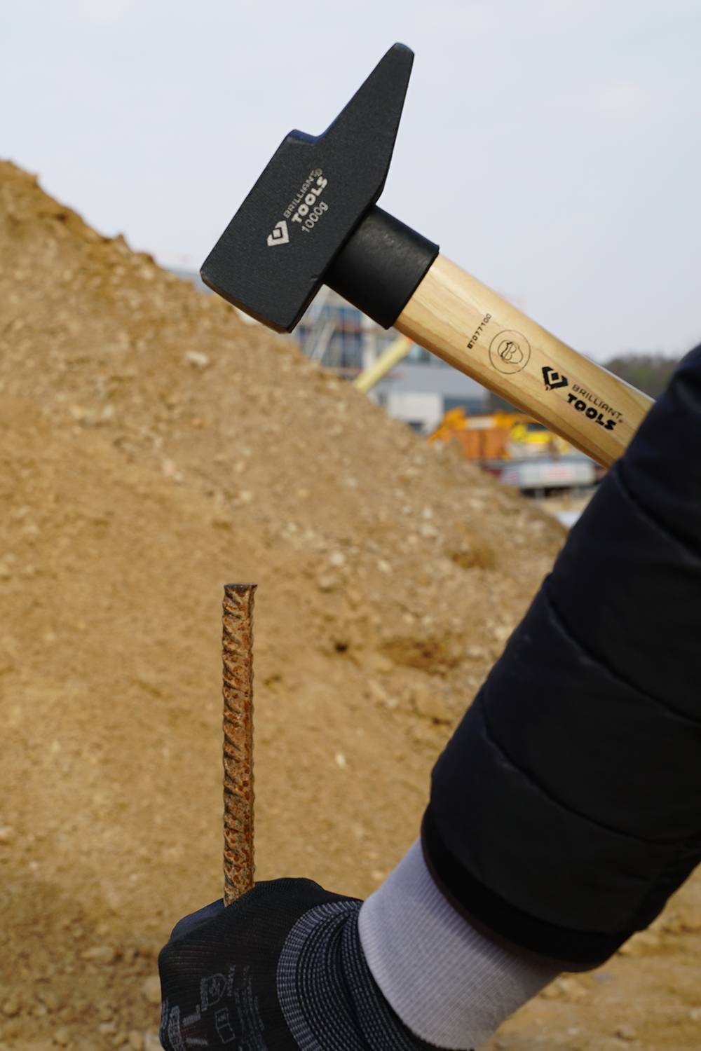 A gloved hand holds a hammer, poised to drive a steel rod into an earth mound. Construction site in the background.