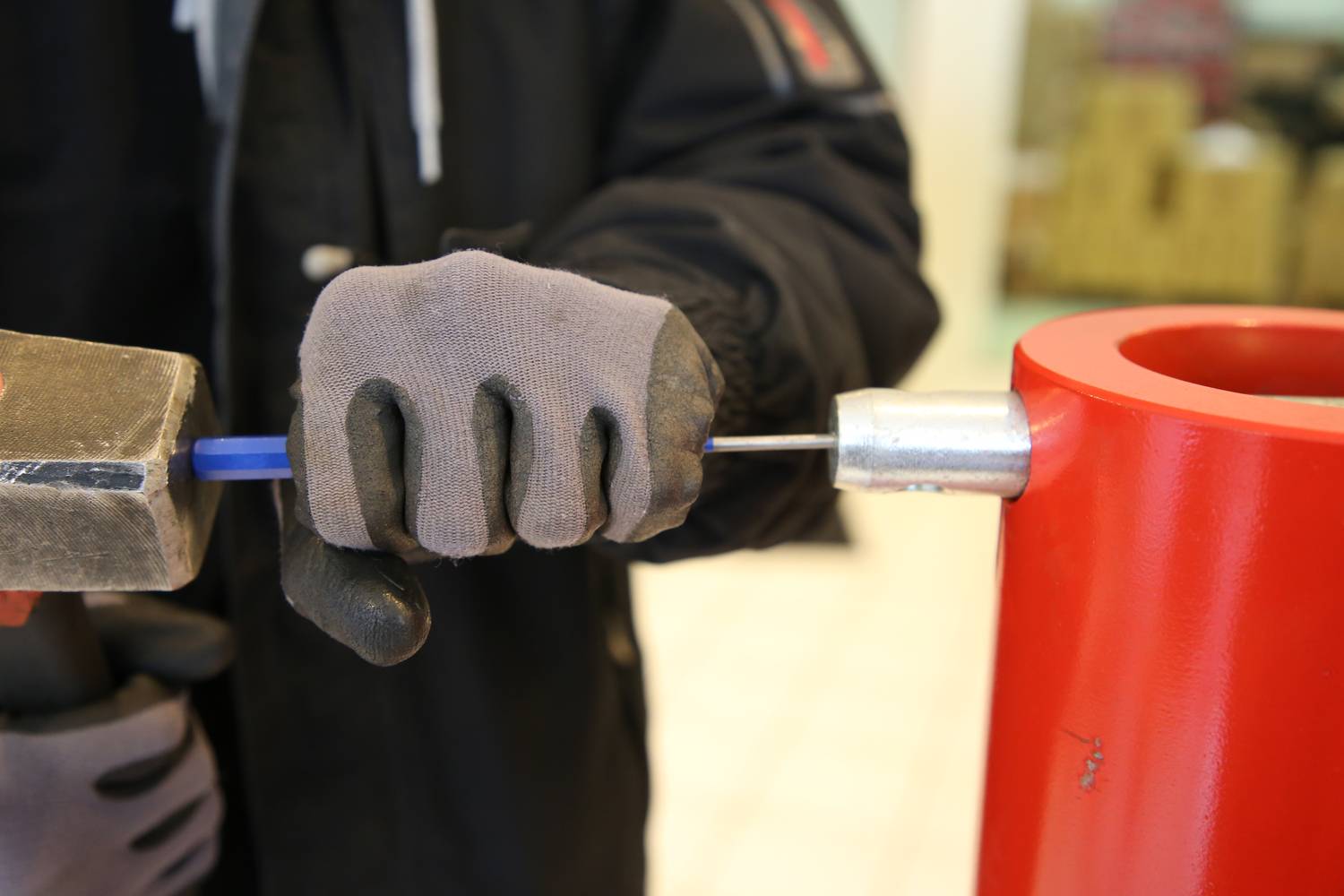 A person is securing a metal element to a blue rod that is inserted into a red metal part using a hammer.