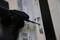A person wearing a black glove is tightening a screw on a hinge, next to a label with text.