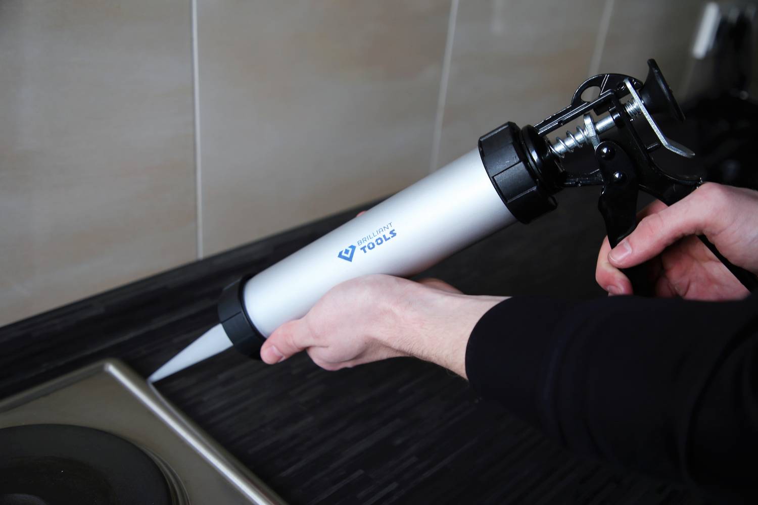 A person is holding a cartridge gun and applying sealant to the joint between the hob and worktop.