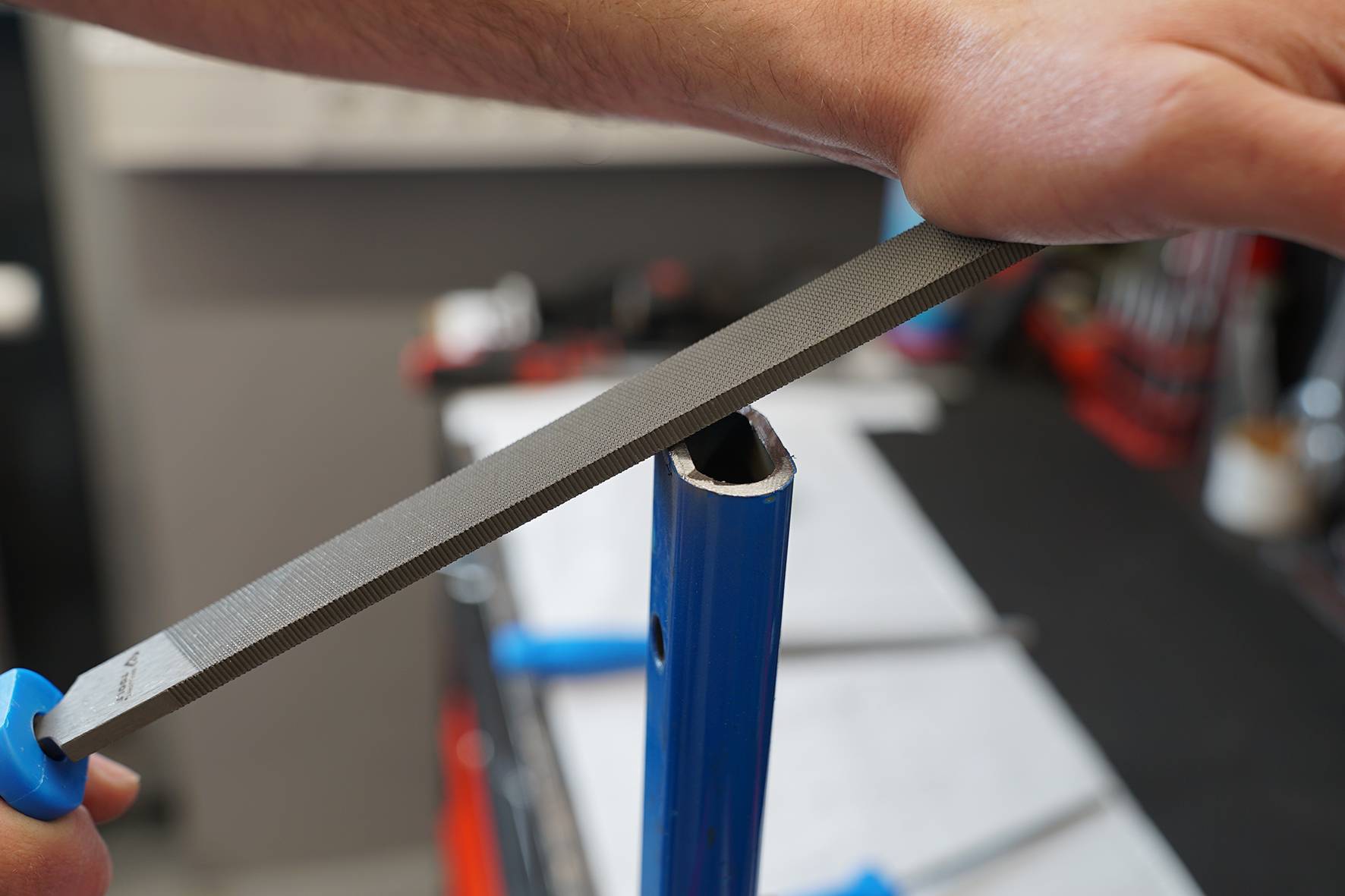 A person is filing the edge of a blue metal pipe with a hand file. Hands and tools are in focus.