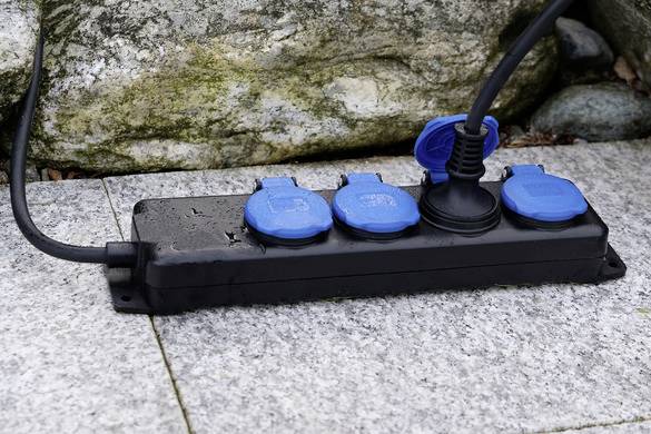 A black multi-socket extension with four blue plug sockets, one of which is in use. It is resting on a grey stone surface.