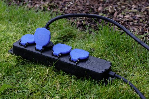 A black power strip with blue covers and an attached cable is lying on wet grass.