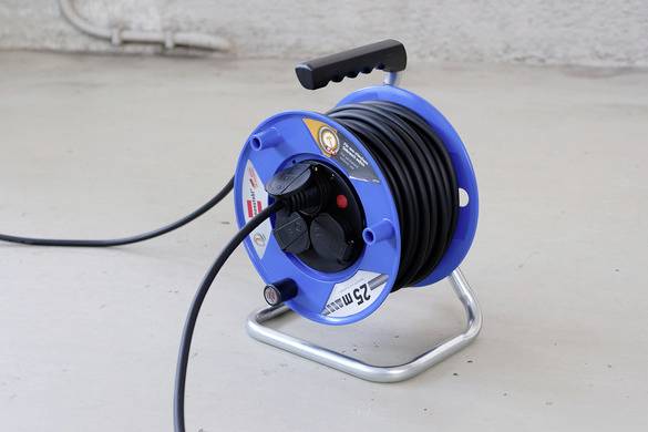 In the picture, a blue cable drum is positioned on a light-coloured floor. A black cable is partially unrolled and plugged into the drum.