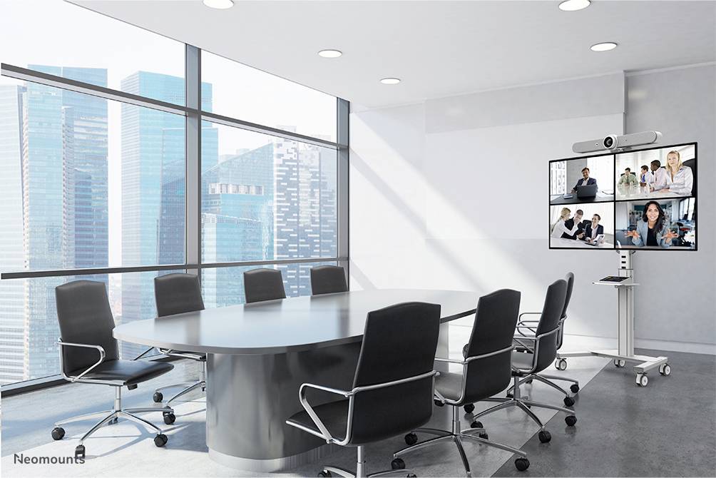 Office conference room with a large oval table and empty chairs. A screen displays a video meeting with multiple participants.