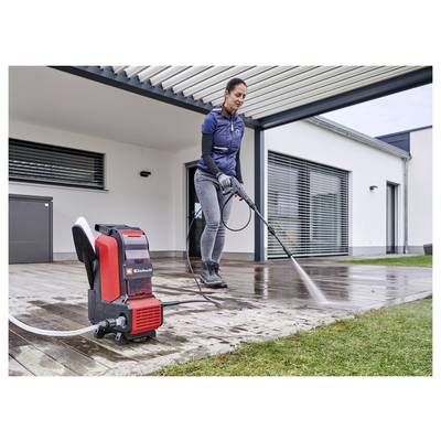 Woman using a pressure washer to clean a wet wooden patio beside a modern house; red portable washer sits on the deck.<br>