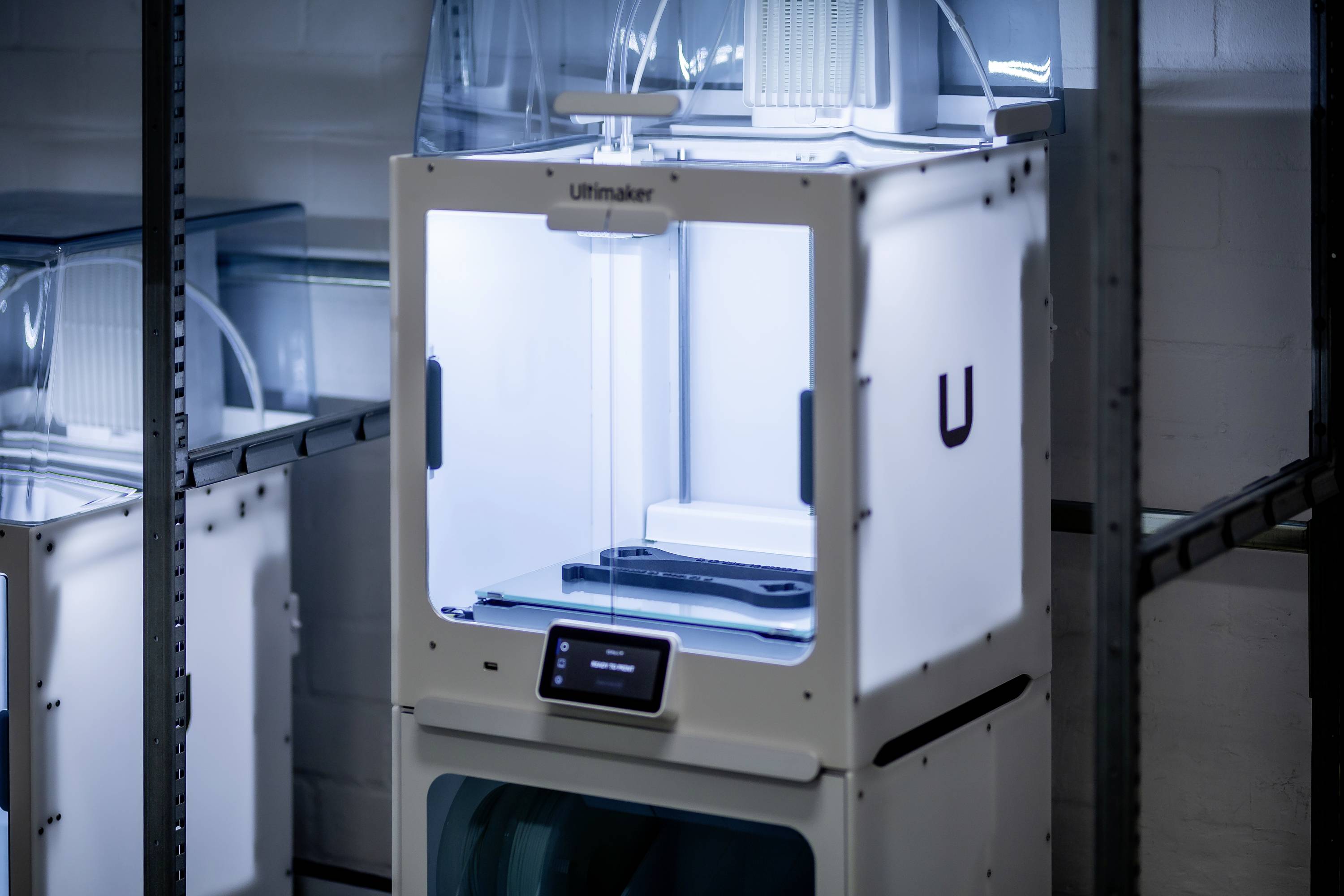 A white 3D printer with an illuminated interior stands on a shelf in a workshop or laboratory environment.