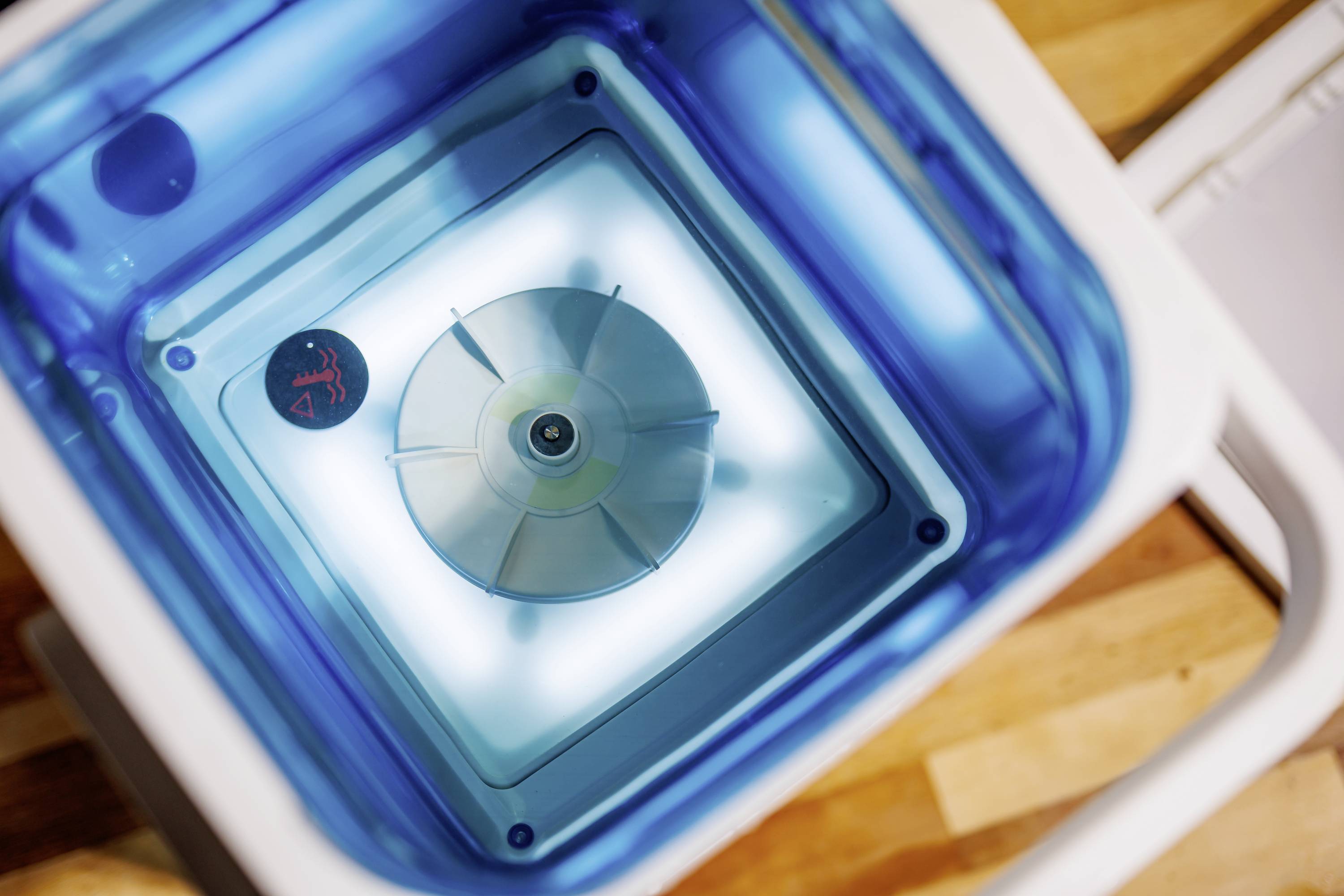 A blue water container viewed from above with a transparent valve in the centre, positioned on a wooden floor.