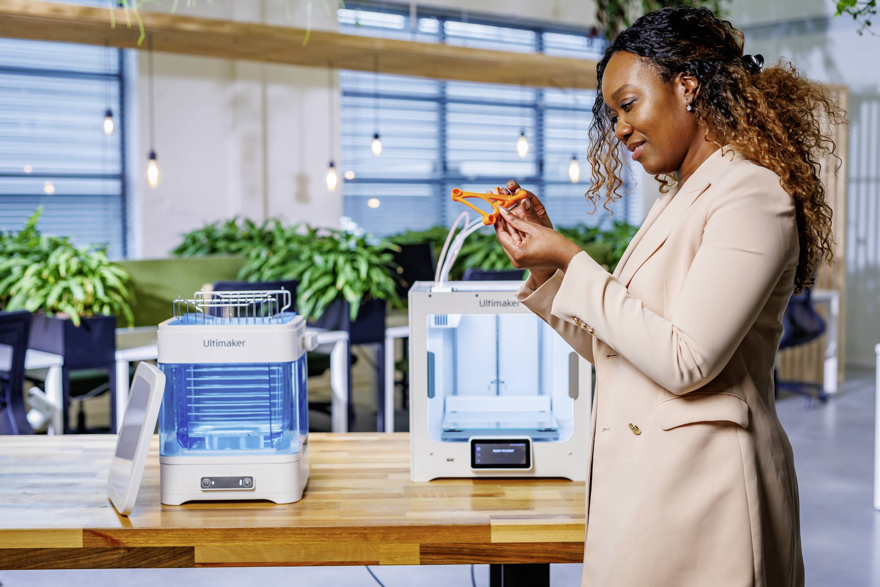 A person is examining an orange 3D-printed object in an office with 3D printers on a table.