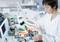 A person in a laboratory is repairing or testing a circuit board with various measuring instruments, surrounded by technical equipment.