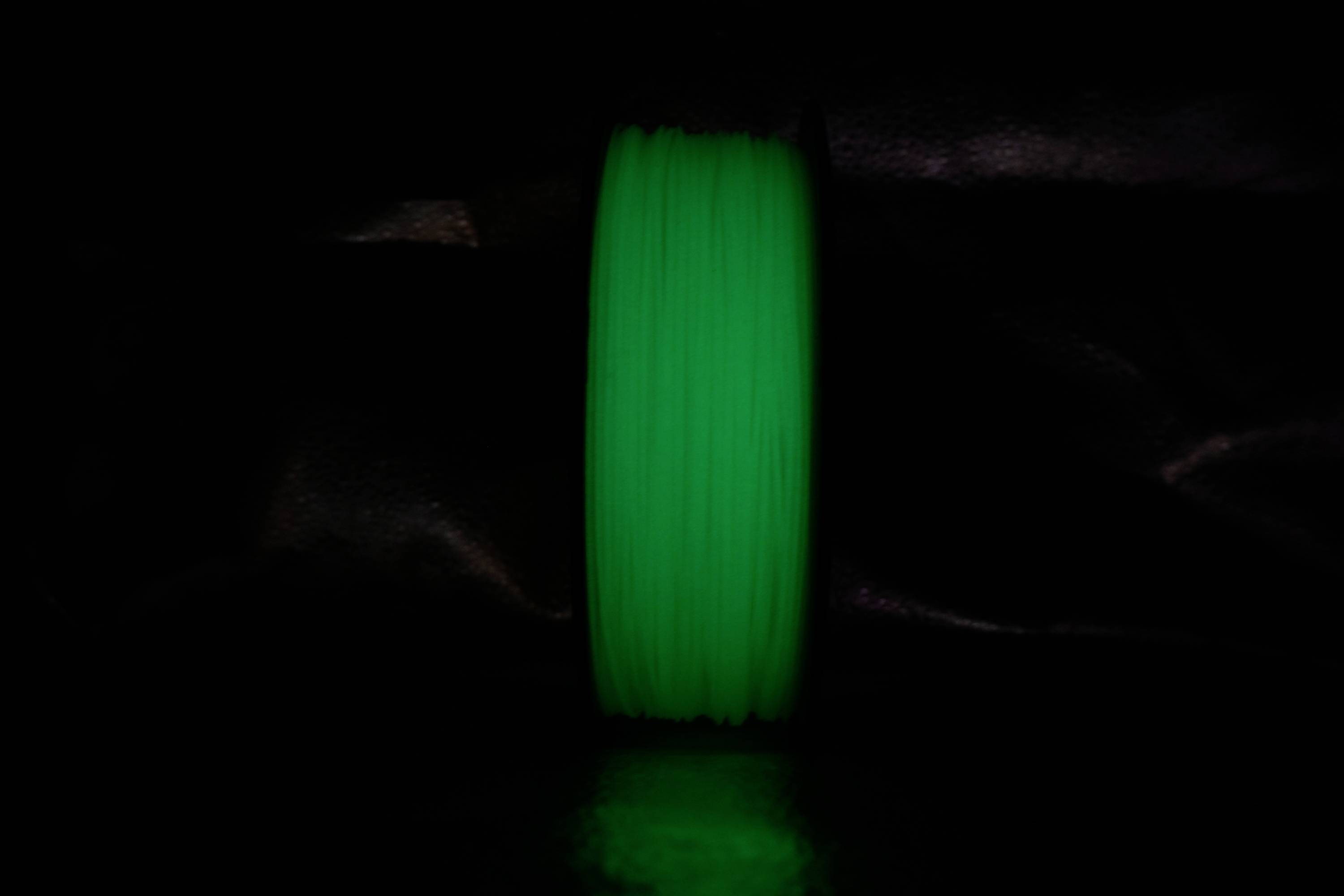 A bright green plastic filament spool in a dark environment. The spool radiates through fluorescent material under low lighting.
