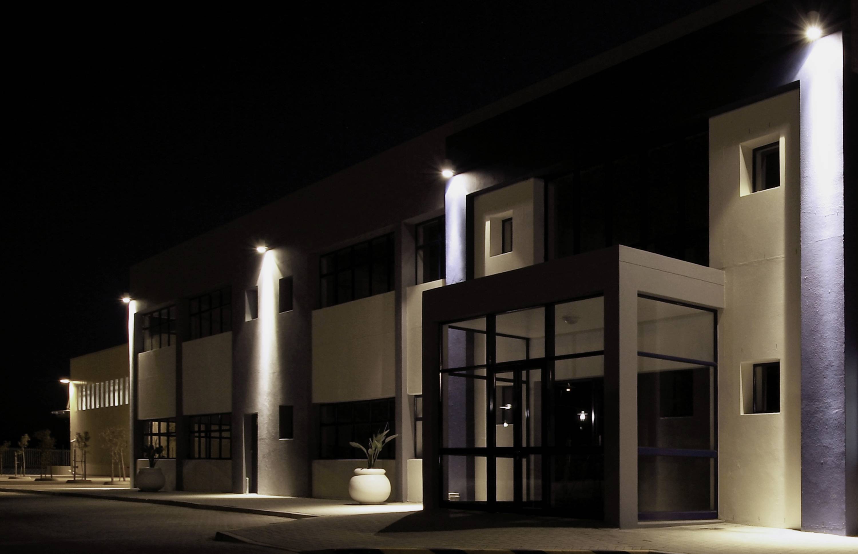 A modern building at night, illuminated by multiple exterior lights, with large windows and a glass entrance area.