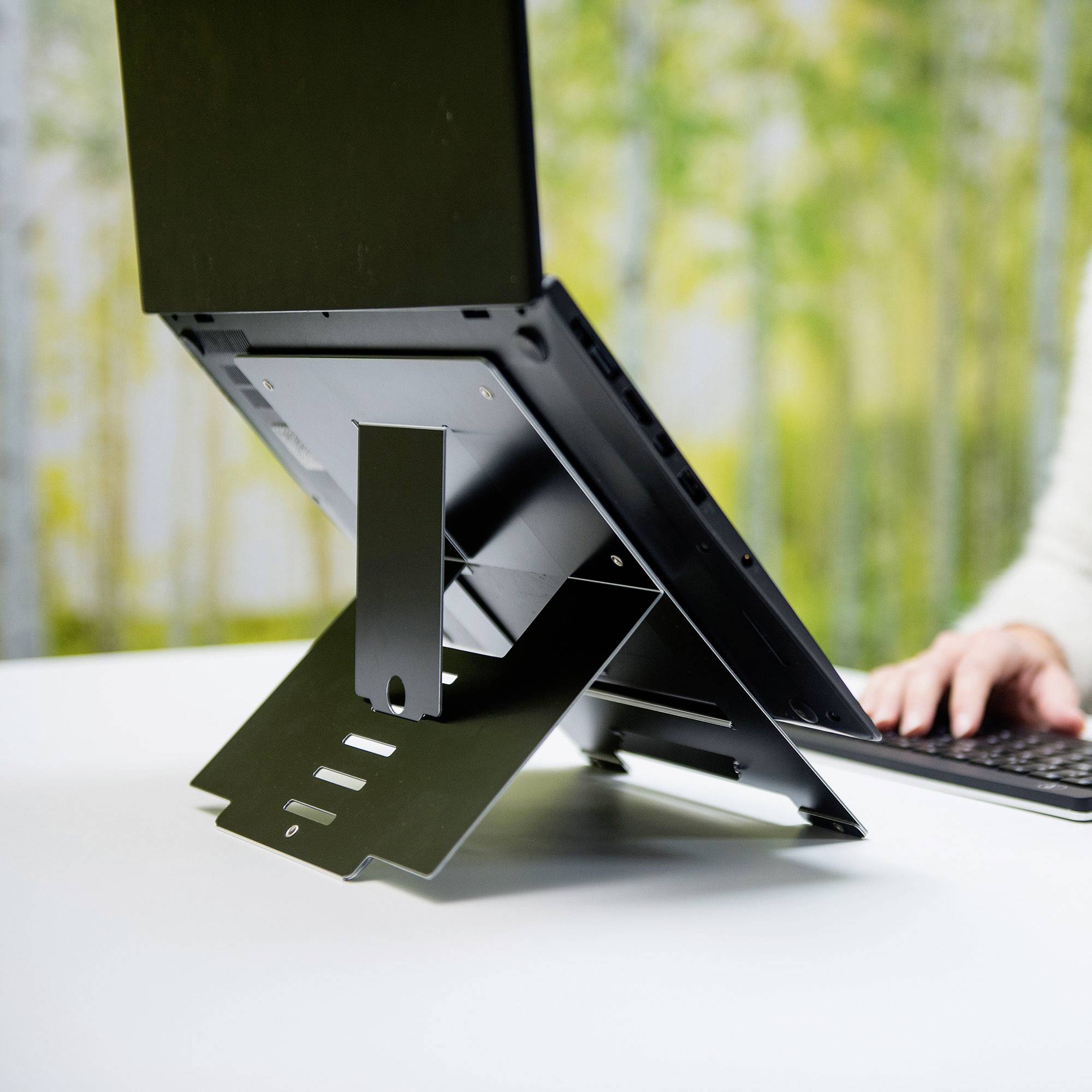 A laptop sits in an adjustable stand on a table. A green plant is blurrily visible in the background.