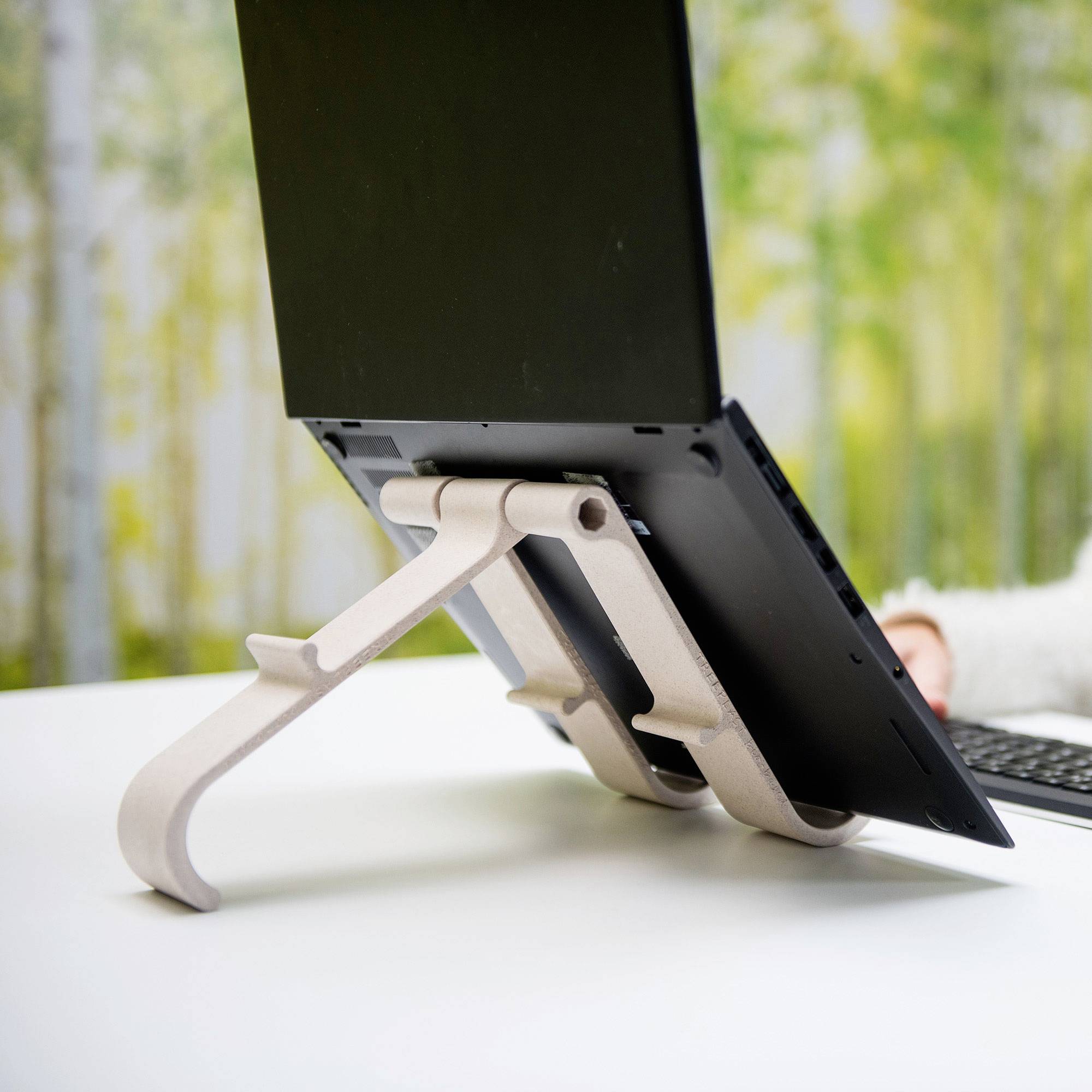 A laptop is sitting on a white table, supported by an angled stand. Trees can be seen blurred in the background.