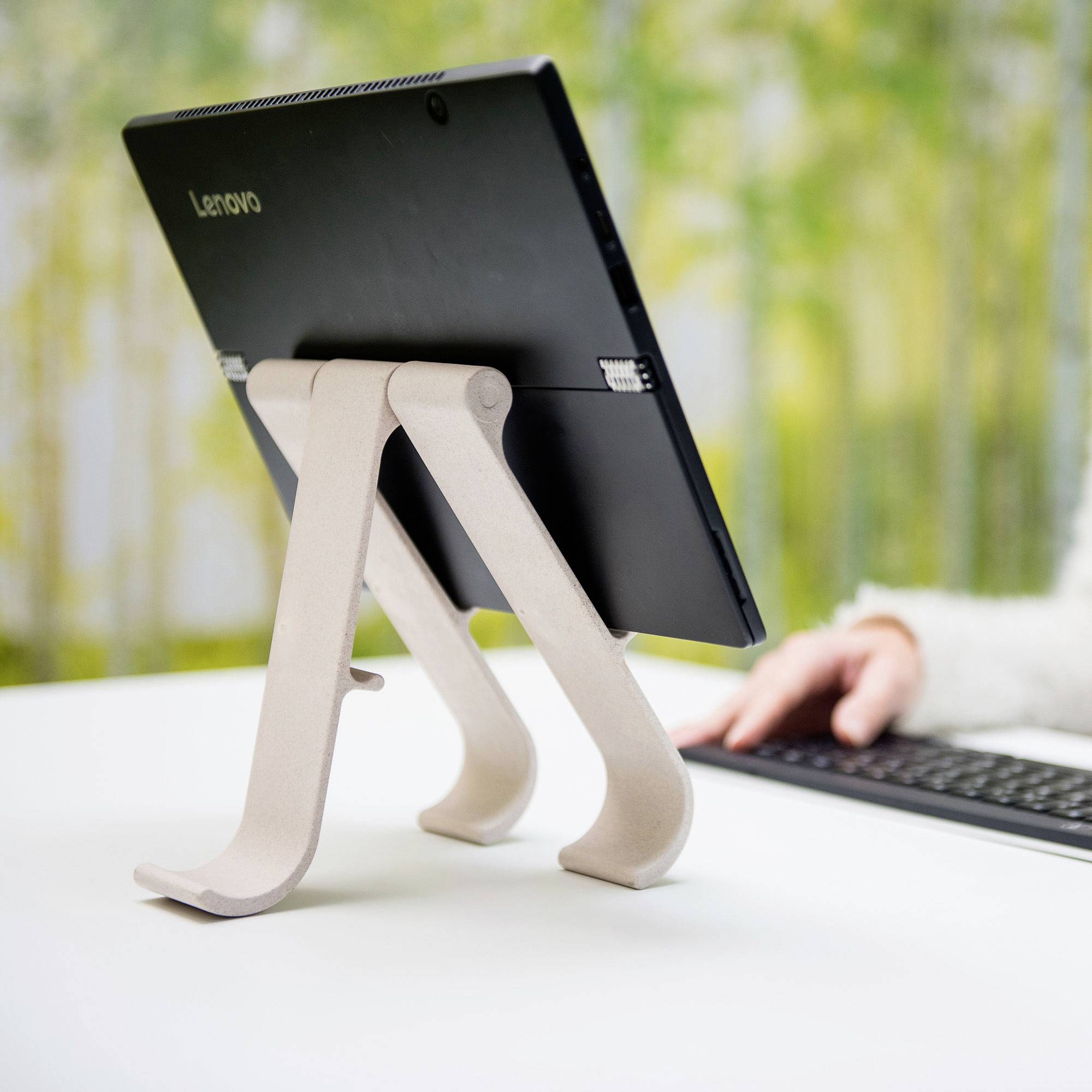 A tablet on a stand, in front of a blurred background. A hand is typing on a keyboard beside it.