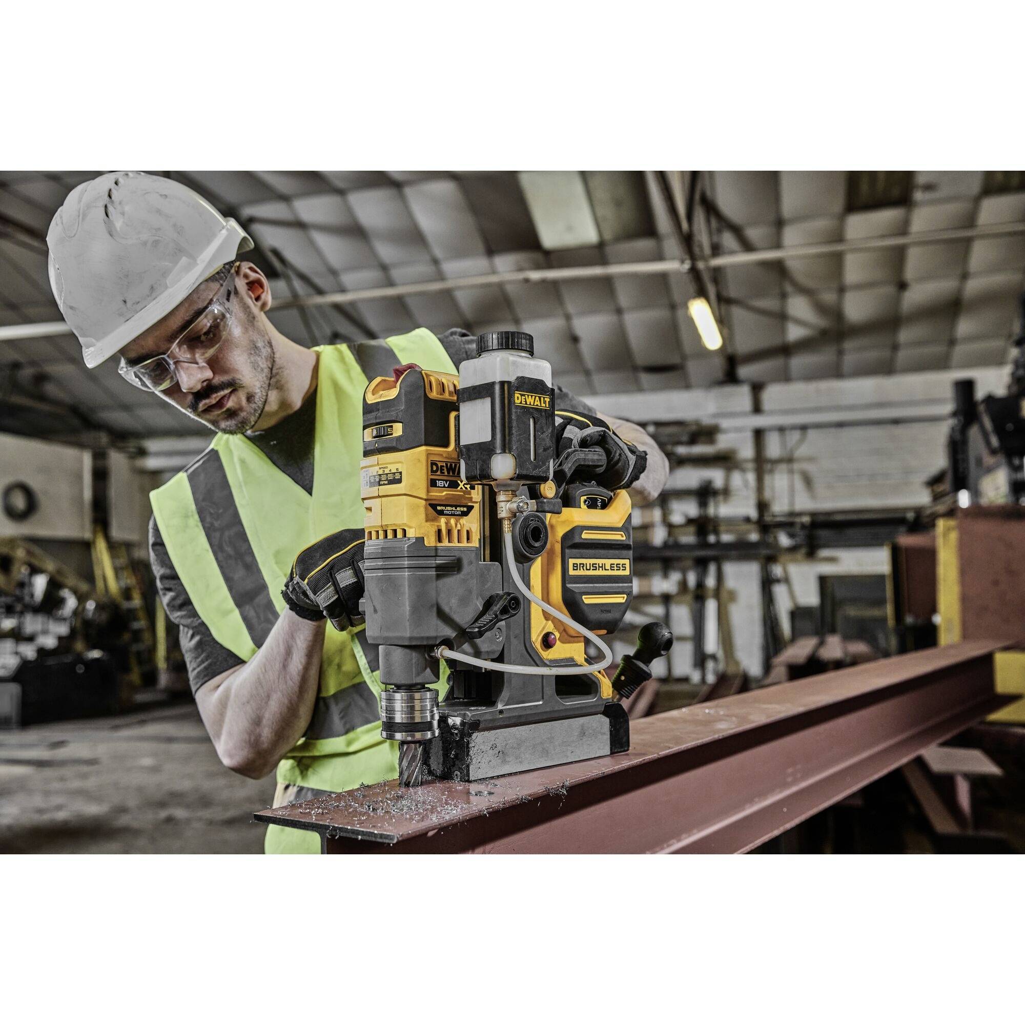 A worker in safety clothing and a hard hat is operating a yellow drill on a steel beam in a workshop environment.