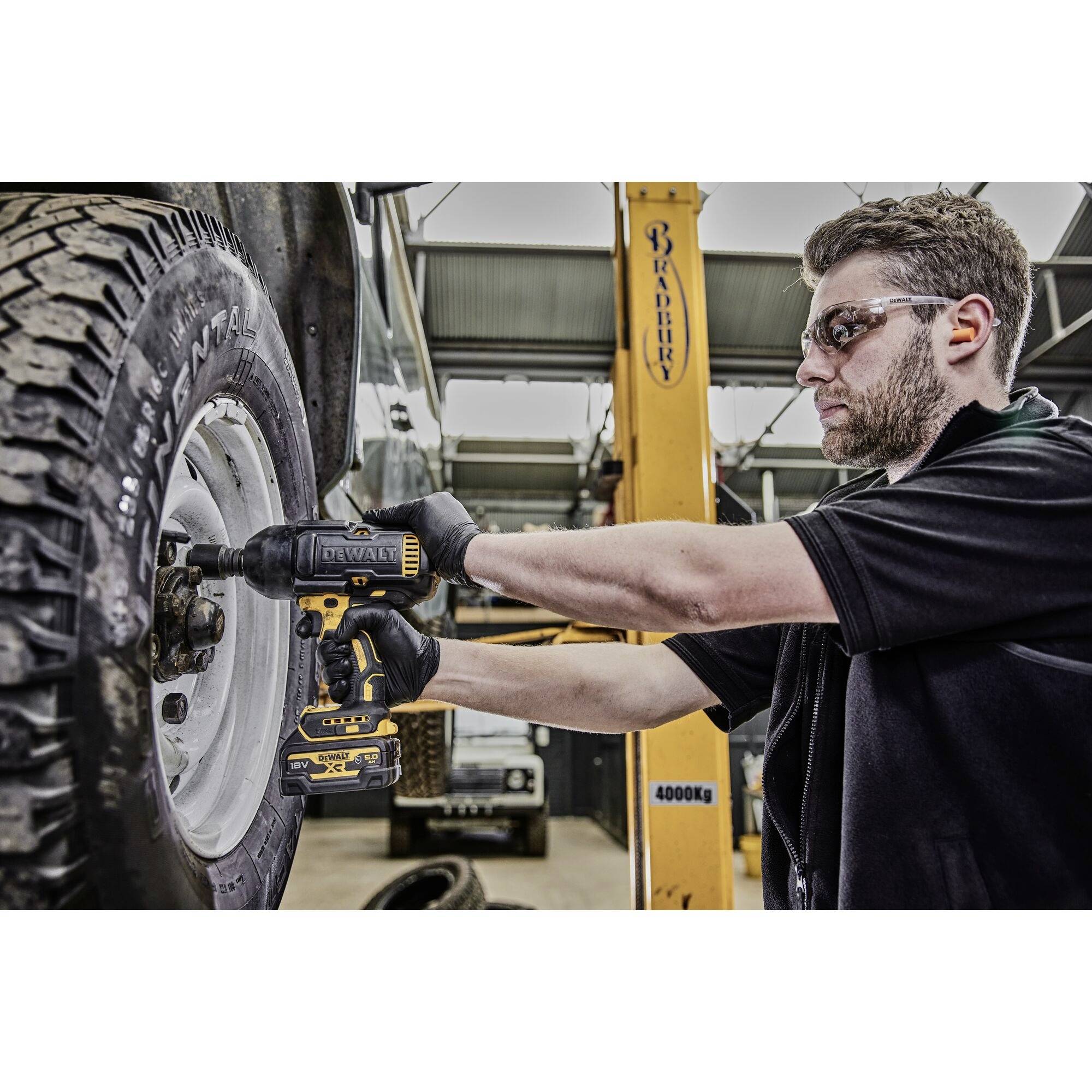 A mechanic wearing safety glasses is tightening the wheel nuts of an off-road vehicle with an electric screwdriver in a workshop.