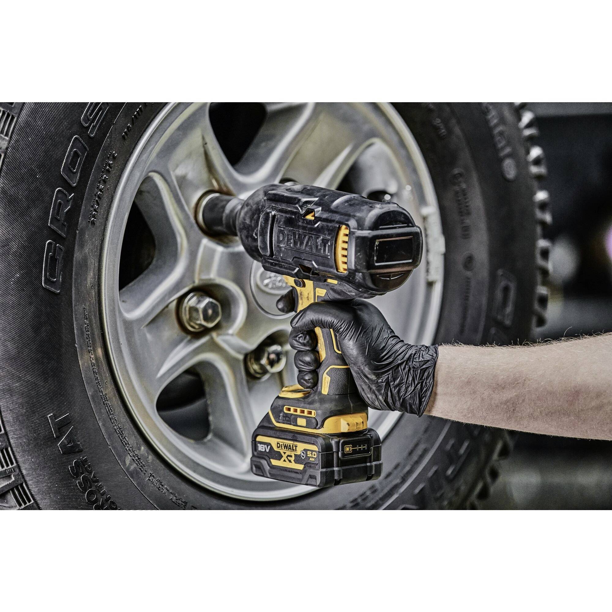 'Right hand wearing a black glove holds a battery-powered impact driver and tightens a wheel nut on the rim of a car.'